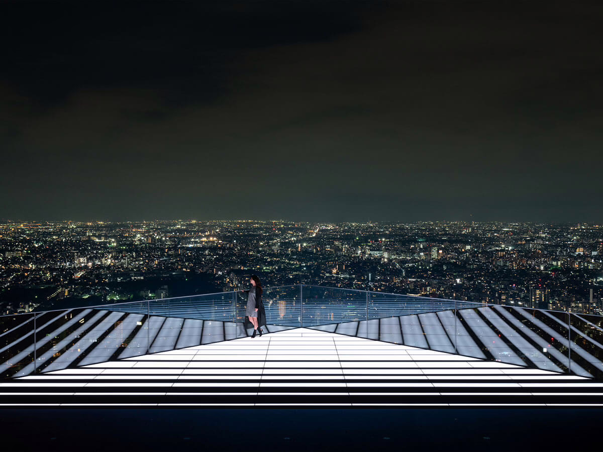 A woman stands on a rooftop observation deck, overlooking a vibrant city skyline illuminated at night.