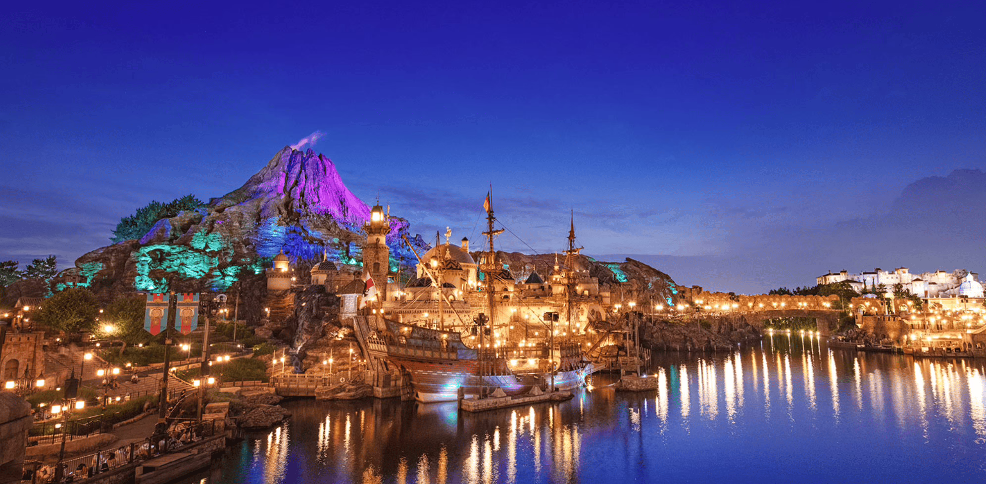 A vibrant theme park landscape with a majestic volcano and illuminated pirate ships reflecting on tranquil waters at dusk.