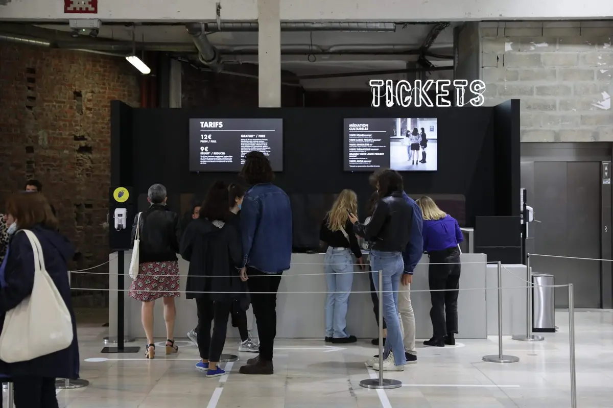 Visitors waiting in line at a ticket counter displaying prices and event information in an arts venue.