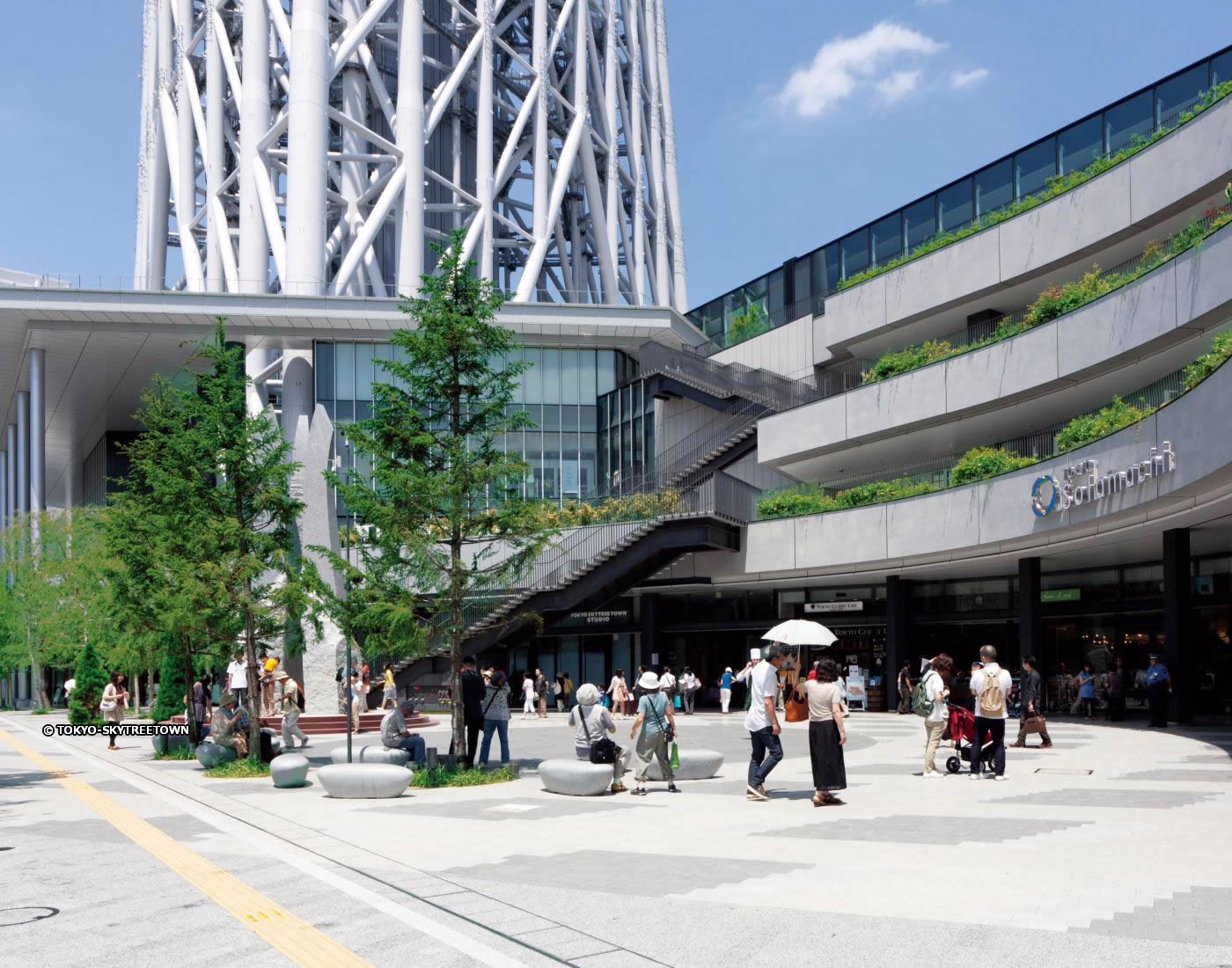 Crowd of visitors enjoying a sunny day at Tokyo Skytree Town, exploring shops and green spaces in a modern urban setting.