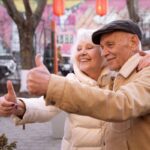 Happy elderly couple enjoying a moment together, both giving thumbs up in a vibrant outdoor setting showcasing joy and companionship.