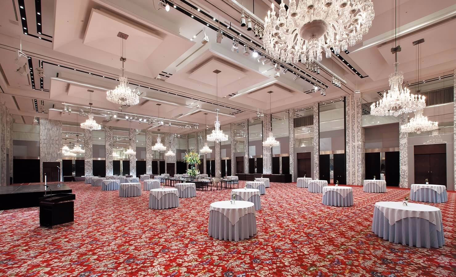 Elegant event space featuring round tables with floral tablecloths, crystal chandeliers, and a vibrant red carpet for gatherings.