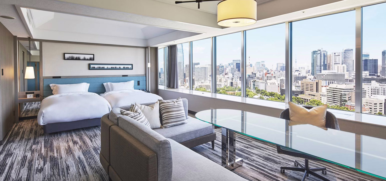 Spacious hotel room featuring twin beds, modern furnishings, and stunning city views through large floor-to-ceiling windows.