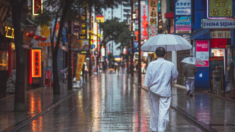 Man in a white outfit walks under an umbrella along a rain-soaked street, surrounded by vibrant neon signs and shops.