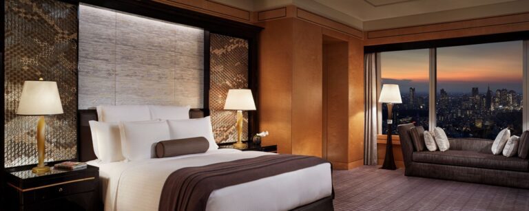 Luxurious hotel room with a king-size bed, elegant decor, and stunning city skyline views at sunset.