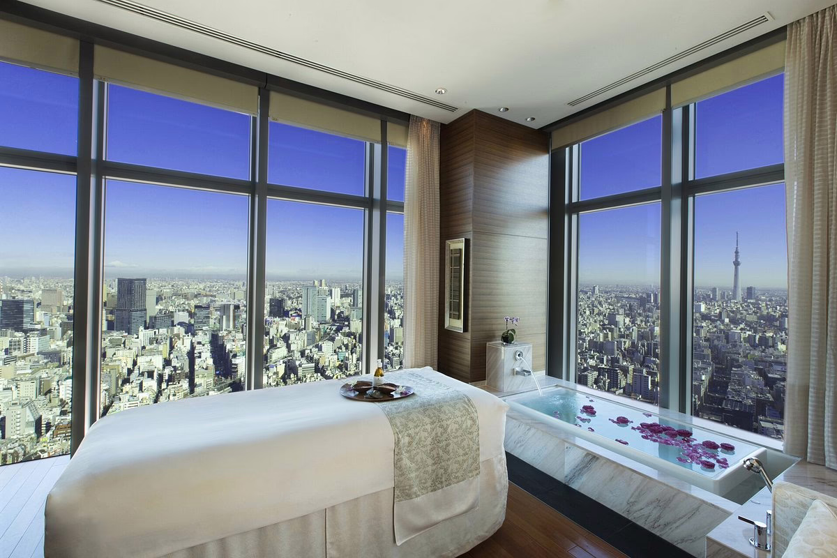 Luxurious spa room with a stunning city view, featuring a massage table and a bathtub adorned with rose petals.