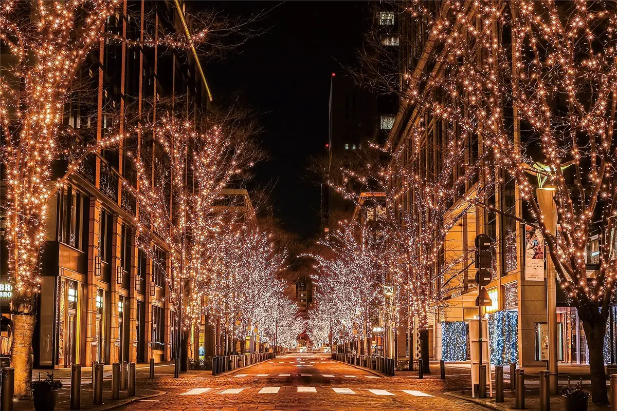 Festively lit city street at night, showcasing glowing trees and vibrant storefronts, creating a warm holiday atmosphere.