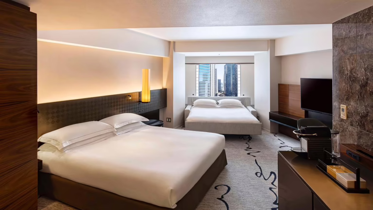 Contemporary hotel room featuring two comfortable beds, stylish decor, and a city skyline view through large windows.