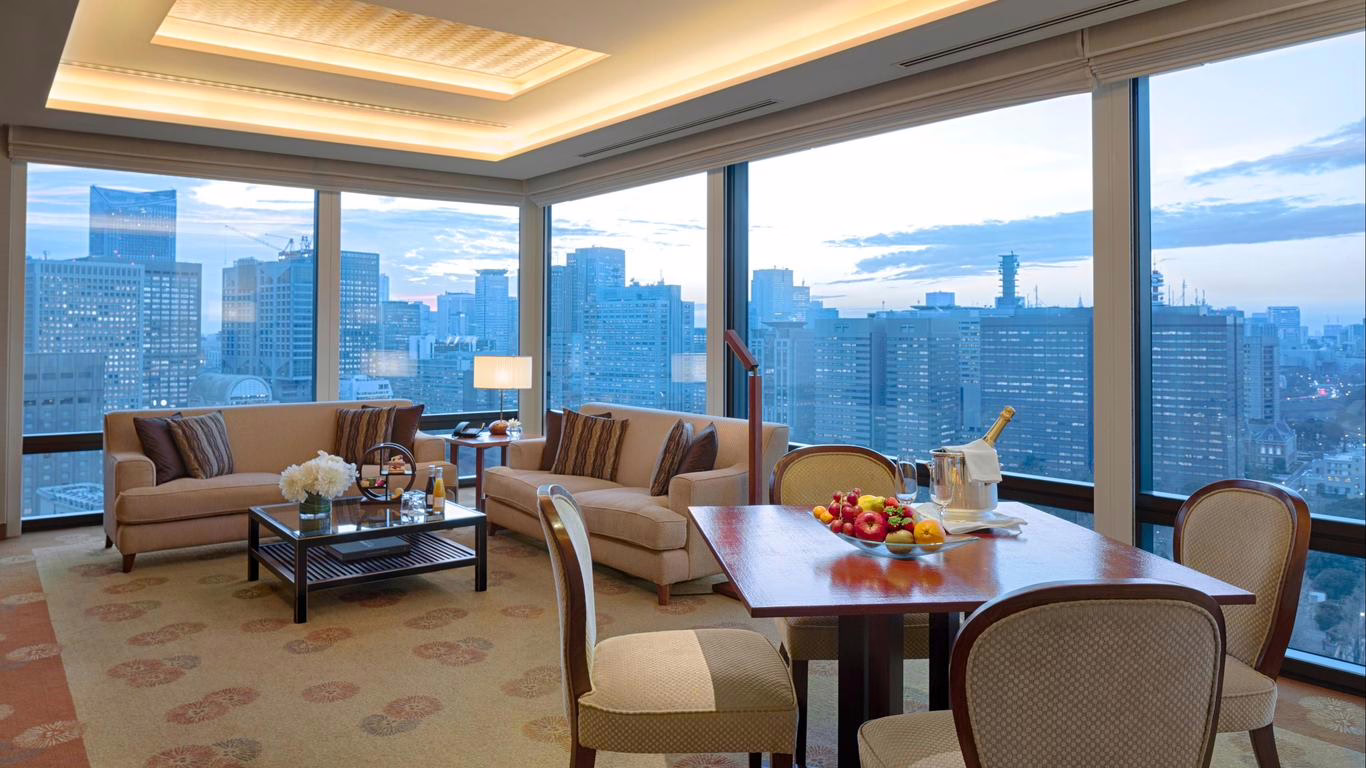 Luxurious hotel suite with a panoramic city view, featuring elegant furnishings and a cozy dining area set for relaxation.