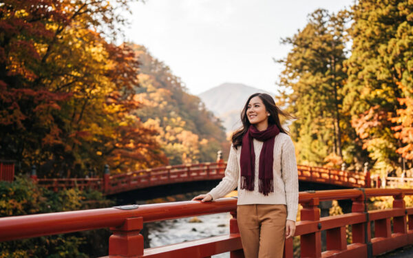 Kannaya Nareswari in a cream sweater and burgundy scarf stands on a red bridge surrounded by autumn foliage, smiling gently.