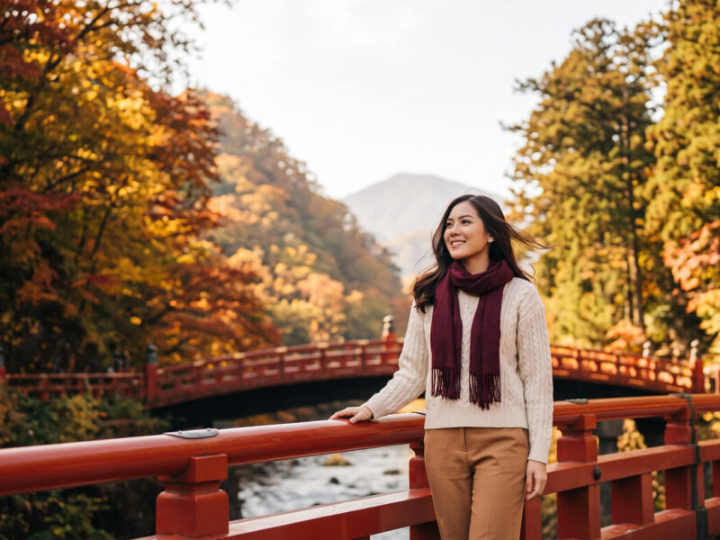 Kannaya Nareswari in a cream sweater and burgundy scarf stands on a red bridge surrounded by autumn foliage, smiling gently.