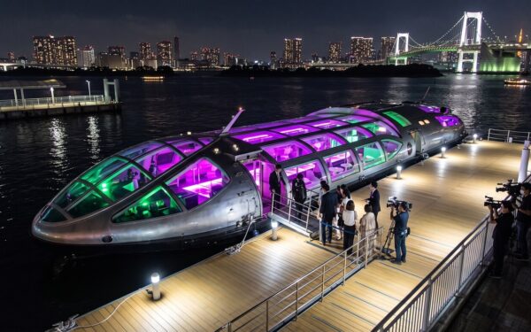 A sleek, futuristic ferry with vibrant purple and green lights docked at night, with people boarding and a film crew present.