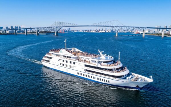 Symphony Moderna Tokyo cruise ship filled with people sails on a bright blue ocean toward a city skyline and a prominent bridge.