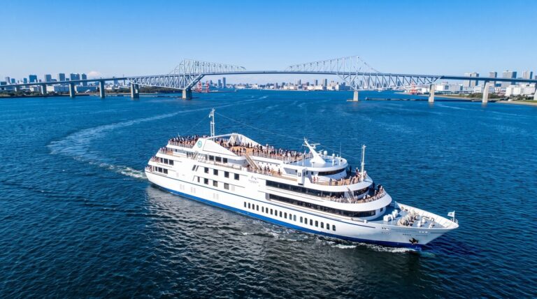 Symphony Moderna Tokyo cruise ship filled with people sails on a bright blue ocean toward a city skyline and a prominent bridge.