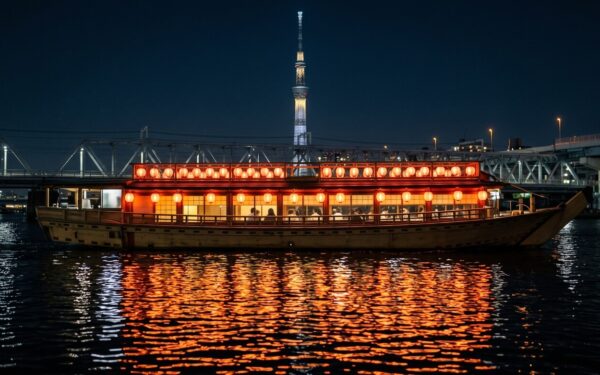 A traditional Japanese boat with red lanterns glides on the water at night, with the Tokyo Skytree visible in the background.